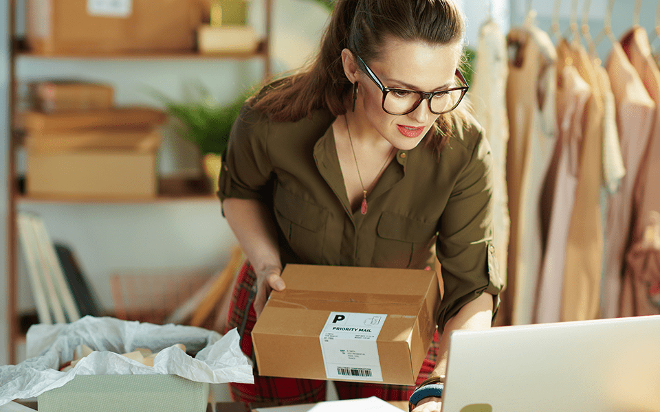 A woman in her office is in front of her computer with a small cardboard box package in her hand