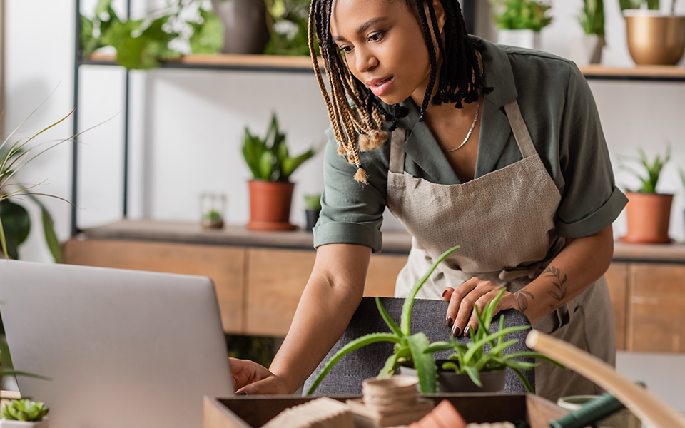 A woman in an office with many plants behind her is leaning toward her desktop computer looking at the screen