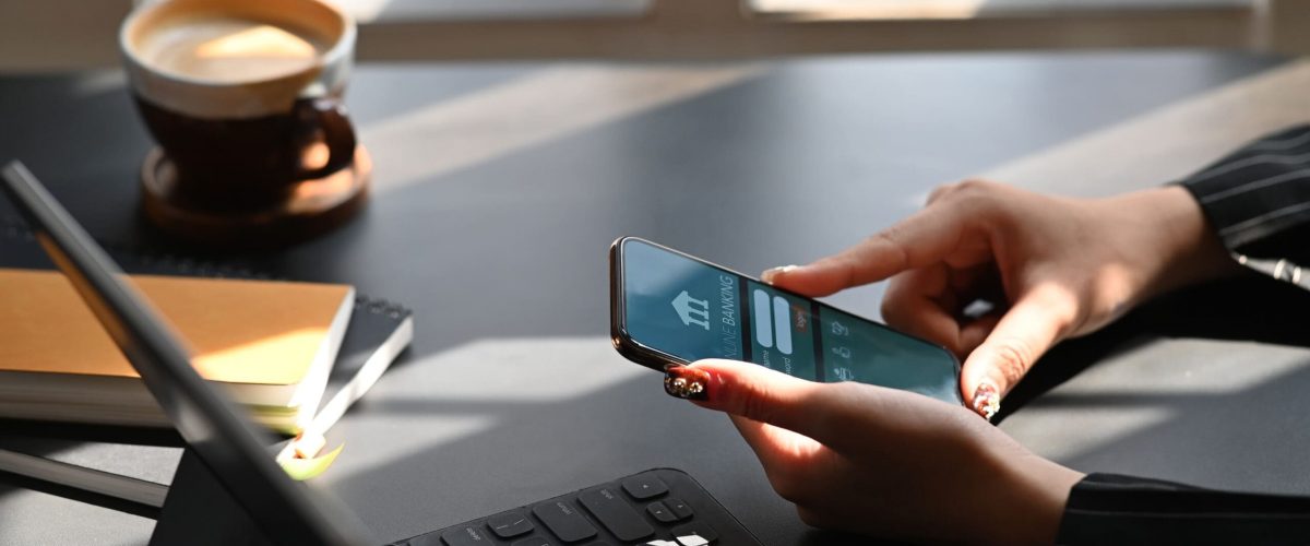 Cropped shot of executive woman holding smartphone in hands while doing online financial transaction by her smartphone in front computer tablet at the modern working desk
