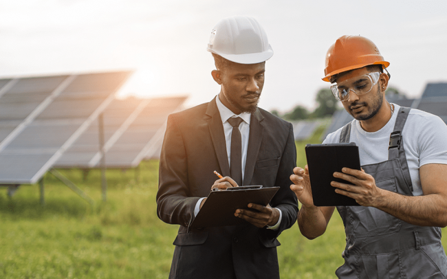 A man in a suit is working outdoor in front of solar panels with another man. Both are wearing hard hats.