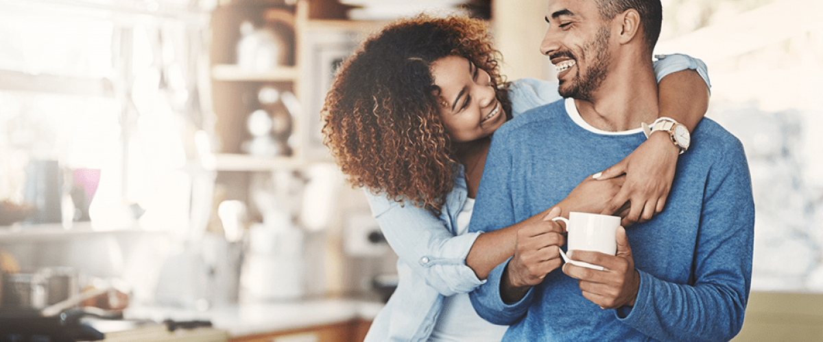A couple is in the kitchen. The man is holding a cup of coffee and his wife has her arms wrapped around his neck, looking at his face lovingly