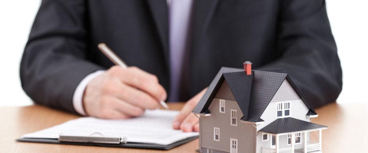 Photo of a man in a suit signing paper work. There is a model of a home in front of it